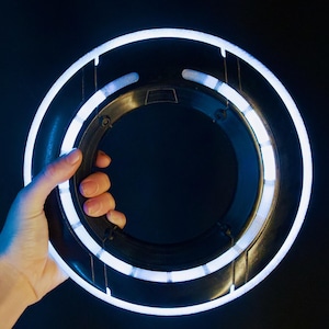 May include: A circular black disc featuring a bright white glowing ring along its outer edge and a smaller inner ring. The object has a smooth, dark plastic frame with structural supports connecting the two rings, creating a futuristic aesthetic.