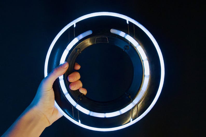 May include: A circular black disc featuring a bright white glowing ring along its outer edge and a smaller inner ring. The object has a smooth, dark plastic frame with structural supports connecting the two rings, creating a futuristic aesthetic.