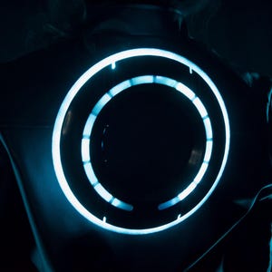 May include: A dark jacket featuring a large, circular, glowing light element on the back. The circular design consists of a continuous outer ring and an inner ring segmented into smaller, illuminated light bars that emit a bright cyan glow.
