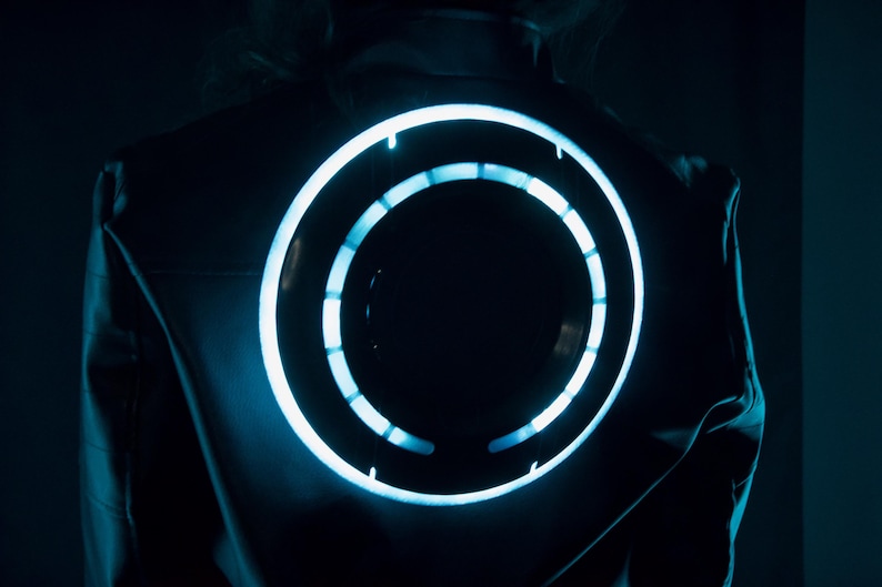 May include: A dark jacket featuring a large, circular, glowing light element on the back. The circular design consists of a continuous outer ring and an inner ring segmented into smaller, illuminated light bars that emit a bright cyan glow.