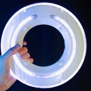 May include: A circular white plastic ring light featuring an integrated glowing LED tube. The device has a hollow center and a smooth, matte finish on the outer frame, emitting a bright, cool-toned white light that illuminates the surrounding area.