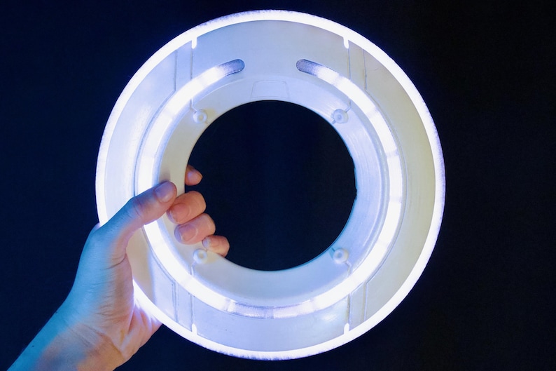 May include: A circular white plastic ring light featuring an integrated glowing LED tube. The device has a hollow center and a smooth, matte finish on the outer frame, emitting a bright, cool-toned white light that illuminates the surrounding area.