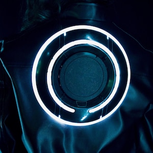 May include: A black jacket featuring a large, circular, glowing light blue neon ring on the back. The design consists of a central dark disc surrounded by a bright, illuminated circular frame that emits a cool-toned light against the dark fabric.