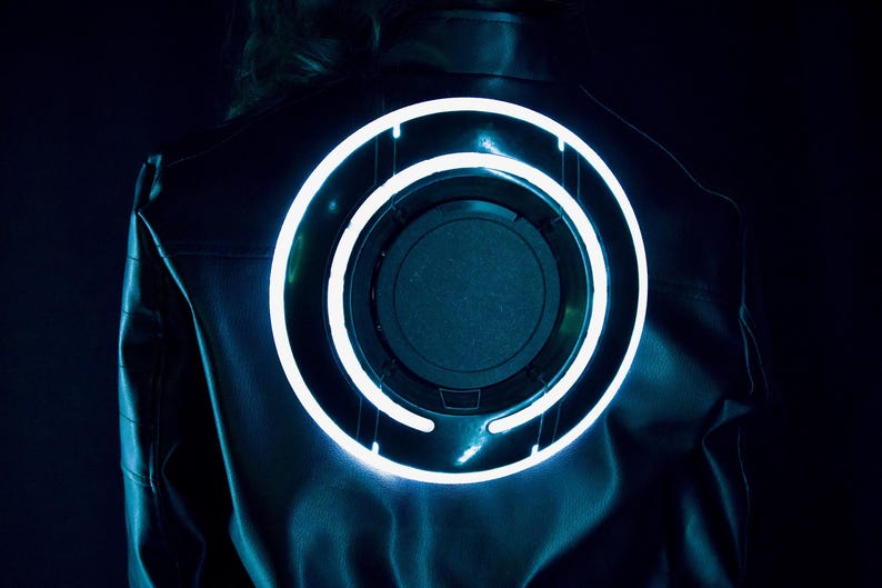 May include: A black jacket featuring a large, circular, glowing light blue neon ring on the back. The design consists of a central dark disc surrounded by a bright, illuminated circular frame that emits a cool-toned light against the dark fabric.