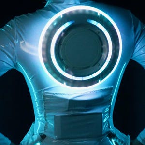 May include: A long-sleeved white bodysuit with a glossy finish featuring a large, circular, glowing blue light disc centered on the back. The garment includes a rectangular panel detail below the disc, creating a futuristic, illuminated aesthetic.
