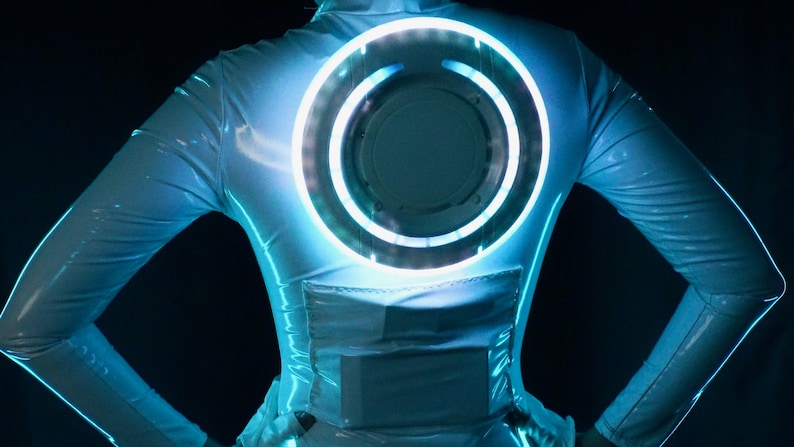 May include: A long-sleeved white bodysuit with a glossy finish featuring a large, circular, glowing blue light disc centered on the back. The garment includes a rectangular panel detail below the disc, creating a futuristic, illuminated aesthetic.