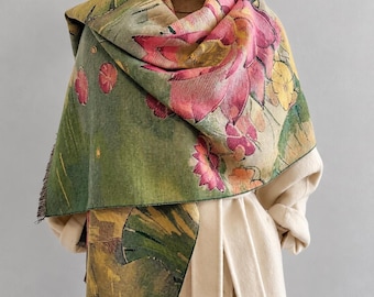 May include: A woven scarf featuring a vibrant floral pattern with large pink lotus flowers, yellow blossoms, and green lily pads on a textured green background. The scarf has a fringed edge and is draped over a beige turtleneck and coat.