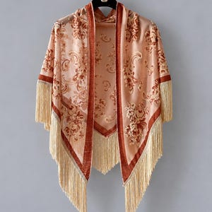 May include: A peach-colored open-front shawl featuring a dark rust-colored velvet trim and a repeating floral paisley pattern. The garment is finished with long, cream-colored fringe along the bottom and sleeve edges, hanging from a black hanger.
