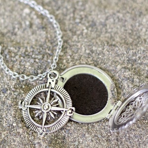 May include: Silver-colored compass pendant necklace with an open locket. The compass design features a detailed starburst pattern. The necklace is on a chain, and the locket is open, revealing a dark interior.