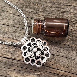 May include: A silver necklace with a beehive pendant featuring a detailed bee design. The necklace comes with a small brown glass bottle.
