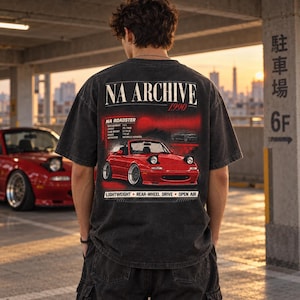 May include: A charcoal grey t-shirt featuring a graphic print on the back. The design displays a red NA Roadster car, the text NA ARCHIVE 1990, and technical specifications including displacement, layout, curb weight, power, and aspiration.
