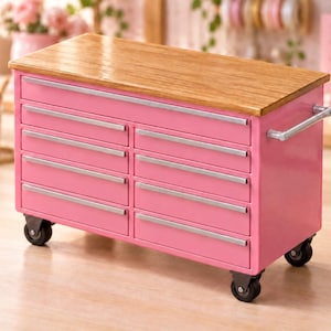 May include: A bright pink metal tool chest featuring ten drawers with silver-toned horizontal handles, a smooth light brown wooden top surface, and four black swivel caster wheels attached to the base for mobility, including a silver side handle.