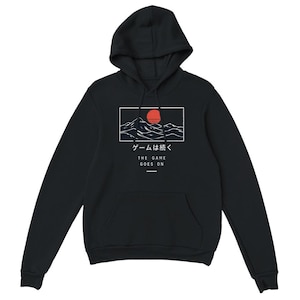 May include: A black long-sleeve hooded sweatshirt featuring a graphic print of a red sun over white mountain outlines. Below the graphic, the text reads "ゲームは続く" and "THE GAME GOES ON" in white lettering, with a small horizontal line underneath.