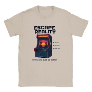 Op de afbeelding: Een beige t-shirt met korte mouwen en een afbeelding van een donkerblauwe arcadekast met een zonsondergang en palmbomen op het scherm. De tekst luidt ESCAPE REALITY, PLAY, RELAX, REWIND en SOMEWHERE ELSE IS BETTER in een pixelstijl.