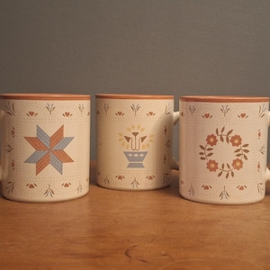 May include: Three cream-colored ceramic mugs with a tan rim. Each mug features a distinct pattern: a blue and orange star, a stylized flower pot, and a circular floral wreath. The designs are framed by small dots and delicate leaf motifs.
