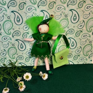 Könnte beinhalten: Eine grüne Filz-Feenpuppe mit einem grünen Filzkleid, goldenen Verzierungen und einer grünen Filztasche. Die Puppe hat braunes Garnhaar und einen grünen Filzhut mit einer Glocke. Die Puppe steht auf einer grünen Filzoberfläche.