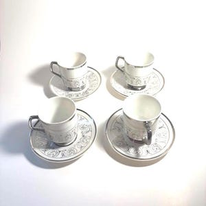 May include: A set of four white porcelain espresso cups and matching saucers, each featuring a silver metallic rim and a repeating silver botanical pattern around the base of the cup and the outer edge of the saucer, with silver handles.