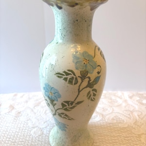 Puede incluir: Un jarrón de cerámica con base de color crema, acabado moteado y diseño floral pintado a mano. El jarrón muestra flores de color azul claro con hojas y tallos verdes que se enroscan alrededor del cuerpo, con un borde y una base acampanados.
