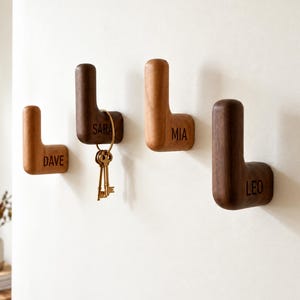 May include: Four L-shaped wooden wall hooks mounted on a white wall. Each hook is engraved with a name: DAVE, SARA, MIA, and LEO. A set of metal keys hangs from the hook labeled SARA. The hooks have a smooth, rounded finish and varying wood tones.
