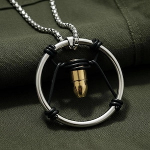 May include: A silver-toned metal necklace featuring a circular pendant frame. A brass-coloured bullet casing is suspended in the centre of the ring by black cord wrappings. The pendant hangs from a silver-toned box chain link necklace.