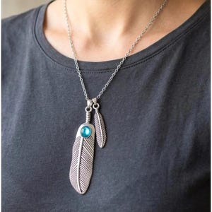 Pode incluir: Um colar de metal prateado com dois pingentes em forma de pena pendurados numa corrente fina. O pingente de pena maior é acentuado com uma única pedra preciosa redonda de cor azul brilhante, fixada no topo da pena de metal texturizada.