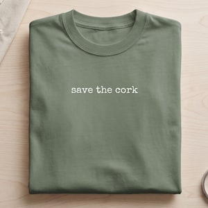 Puede incluir: Una camiseta marrón de cuello redondo con el texto blanco "save the cork" impreso en una fuente estilo máquina de escribir en el centro del pecho. La prenda está doblada cuidadosamente, mostrando las mangas cortas y el cuello acanalado.