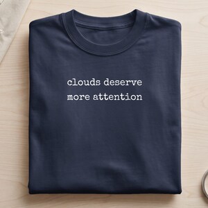 May include: A brown crew neck t-shirt featuring the white text "clouds deserve more attention" printed in a typewriter-style font across the centre of the chest. The garment is folded neatly, showcasing the short sleeves and the ribbed collar neckline.