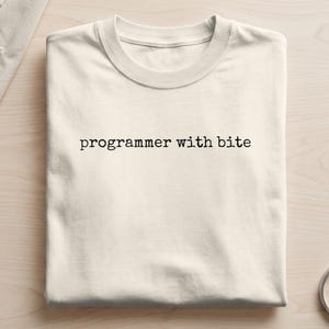 May include: A brown short-sleeved t-shirt featuring the white text "programmer with bite" printed in a typewriter-style font across the chest. The garment has a crew neckline and is folded neatly, displaying a smooth, solid-coloured fabric surface.
