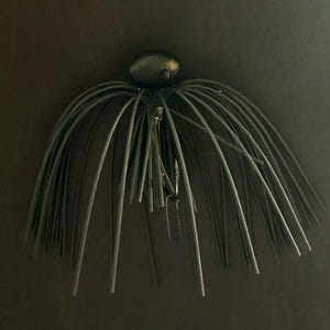 Puede incluir: Un señuelo de pesca negro con una cabeza redondeada y una falda compuesta por numerosos hilos de silicona negros, finos y flexibles. Un anzuelo de metal afilado es parcialmente visible, extendiéndose desde el centro de la cabeza hacia abajo.