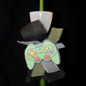 May include: A green candle with a white plastic cup base, decorated with a green video game controller, black mesh, and green and white ribbons. The controller has orange, red, and purple buttons.
