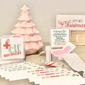 May include: A pink ceramic Christmas tree, a wooden countdown block that says "Sleeps Until Christmas" with the number 6 showing, and a clear plastic container with a red cross and the words "Elf Recovery" on it. The container has a small pink bandage, a roll of white gauze, a small bottle of glitter, and a candy cane inside. There are also several small white cards with the words "An Elf Warning for You" written on them.