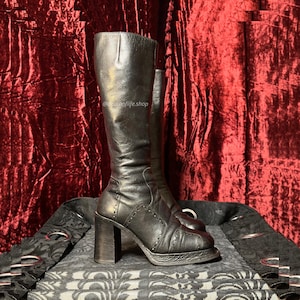 May include: A pair of black knee-high boots featuring a block heel, a textured platform sole, and decorative stitching along the side panels. The boots have a smooth finish and a vertical seam running up the front shaft. The text @deathoflife.shop is visible.