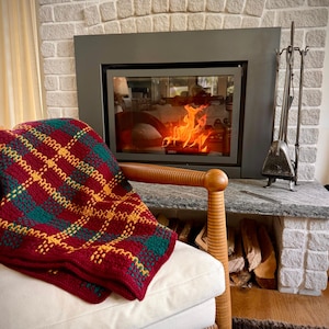 May include: A crocheted throw blanket featuring a plaid pattern in deep red, forest green, and golden yellow yarn. The blanket is draped over a light-colored armchair with a wooden frame, positioned in front of a fireplace with a burning fire.
