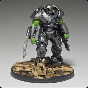 May include: A painted miniature figurine of a green-skinned humanoid creature wearing metallic silver armour. The figure features a curved blade on the right arm, a firearm on the left, and stands on a textured base with a skull-like object.