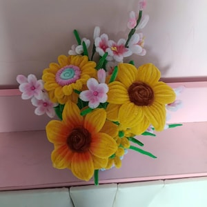 Pode incluir: Um buquê artesanal feito de limpadores de cachimbo felpudos, apresentando três grandes girassóis amarelos com centros castanhos, um girassol com centro azul claro e várias pequenas flores rosa claro com caules e folhas verdes.
