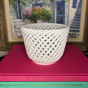 Peut inclure: Un panier en céramique blanche avec une texture rappelant une corde tressée et un motif en treillis diagonal. Le bol présente une ouverture circulaire et une base solide, mettant en valeur un design détaillé imitant des cordages torsadés.