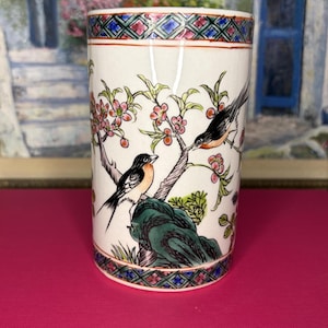 May include: A cylindrical ceramic vase featuring a cream-colored base with hand-painted illustrations of two birds perched on flowering branches above a green rock. The top and bottom rims are decorated with a colorful geometric diamond pattern.