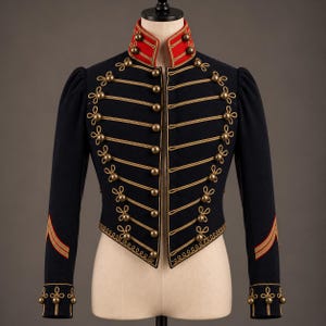 May include: A dark navy blue military-style jacket featuring a high red collar with gold trim, gold-toned buttons, and intricate gold cord braiding across the front. The sleeves include gold cuff details and red and gold stripes near the wrists.