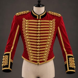 Può includere: Una giacca in stile militare rossa con dettagli in cordoncino dorato intrecciato sul davanti, bottoni color oro, colletto nero e polsini neri. Le spalle sono accentuate da spalline intrecciate dorate e le maniche presentano bordini dorati.