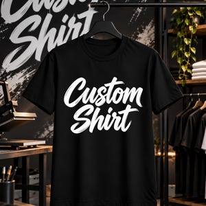 Può includere: Una t-shirt nera a maniche corte appesa a una gruccia nera. La parte anteriore della maglietta presenta la scritta "Custom Shirt" stampata in un carattere bianco in grassetto, in stile pennellata, centrata sul petto.