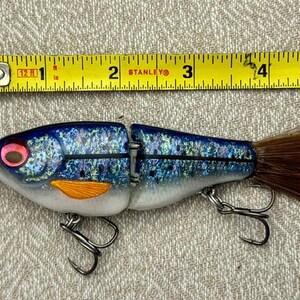 May include: A blue and silver jointed fishing lure with a glittery finish, orange pectoral fin, red eye, and brown feather tail. A yellow Stanley measuring tape positioned above shows the lure is approximately 4.5 inches in length.