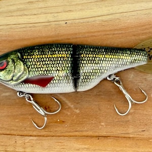 May include: A two-piece jointed fishing lure with a metallic green and silver scale pattern, a red eye, a red pectoral fin accent, and a translucent tan tail with black speckles. The lure features two silver treble hooks attached to the underside.