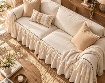 May include: A cream-colored sofa cover with a ruffled lace trim along the bottom edge. The cover features a textured fabric and is styled with two solid tan square throw pillows and one rectangular pillow with striped patterns.