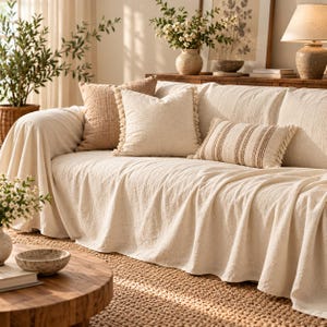 May include: A cream-colored textured sofa cover draped over a couch, accompanied by a square beige pillow with tassel trim, a solid tan pillow, and a rectangular lumbar pillow featuring brown vertical stripes and fringed edges.