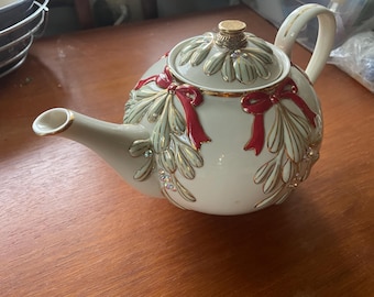 May include: A cream-coloured ceramic teapot featuring a raised mistletoe pattern in muted green and gold, accented with bright red bows and small clear crystals. The teapot includes a matching lid with a gold-tone knob and gold-trimmed edges.
