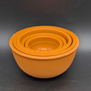 May include: A set of three nested bowls in a solid matte orange finish. The bowls are circular with smooth, rounded edges and a consistent color throughout, stacked one inside the other to create a compact, layered appearance from above.
