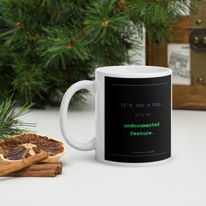 May include: A white ceramic mug featuring a black rectangular graphic with the white text It's not a bug. It's an undocumented feature. and the green text undocumented feature. The bottom includes the text // working as intended.