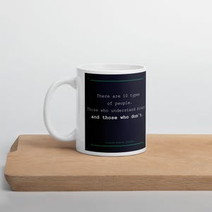 Può includere: Una tazza in ceramica bianca con un pannello rettangolare scuro che riporta il testo: There are 10 types of people. Those who understand binary and those who don't. Il design include sottili linee orizzontali color ottanio sopra e sotto.