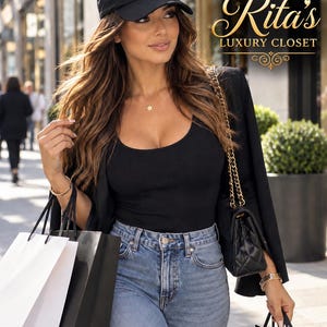May include: A black baseball cap featuring an embroidered eye design with blue, white, and black details and dangling pearl-like accents. The cap is worn with a black scoop-neck top, a black blazer, and blue denim jeans, accompanied by shopping bags.