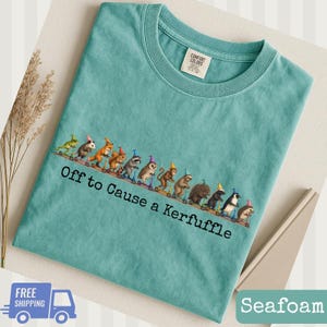 May include: A teal crew neck t-shirt featuring a graphic of various animals wearing party hats and riding scooters in a line. Below the animals, the text reads "Off to Cause a KERFUFFLE" in black lettering. The shirt has a tag at the neck.
